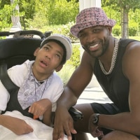 Amari sits in a wheelchair wearing a white T-shirt and white baseball cap. On the right, Tristan Thompson smiles at the camera wearing a pink tweed bucket hat and black tank top.