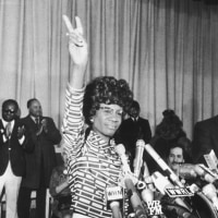 Shirley Chisholm behind microphones holds her right hand in a peace sign.