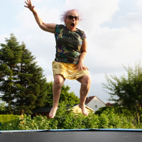 Eccentric senior woman with dyed pink hair, wearing a black shirt with colorful pattern and a golden skirt, jumping on trampoline