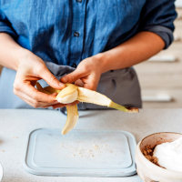 Hands of a woman preparing peeling a banana with yogurt on the side.