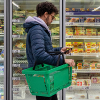 Man shopping in a supermarket while on a budget. He is looking for low prices due to inflation, standing looking at his phone in front of a row of freezers. He is living in the North East of England.