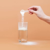 Woman pours creatine powder in a glass of water on a beige background. 