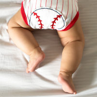 A baby on the bed is wearing a baseball outfit.