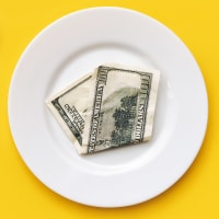 A hundred-dollar bill on a white plate between a fork and a knife on a yellow tablecloth.