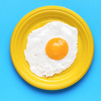 A fried egg in a yellow plate and a yellow fork and spoon on a blue background. A popular breakfast. Minimal concept. A popular breakfast.