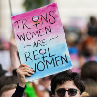 Two people hold signs outside in a crowd, sign on the left says "Trans women are real women", sign on the right says "Science says trans people are real"