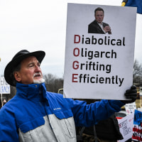 A person holds a sign outside that reads "Diabolical Oligarch Grifting Efficiently"
