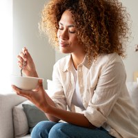 Young relaxed pensive African American woman eating healthy breakfast sitting on the couch.