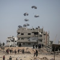 Parachutes fall near a partially destroyed building as people approach