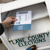 A hand hovers a ballot over a ballot drop box