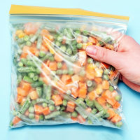 Female hand holding zip bag with frozen vegetables on blue background close-up.