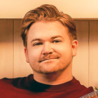 Hunter Hayes sits on chair in a living room holding a guitar, with the fireplace going and Christmas tree lights on