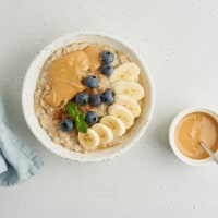 Oatmeal porridge with peanut butter, blueberry, cinnamon, banana. Traditional bunting breakfast served in ceramic bowl on gray background. Top view. Vegetarian and nutritious vitamin morning eating. Classic hot homemade dietary meal