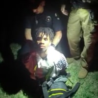 A young black man is held from behind as he kneels in the grass, illuminated by flashlights.