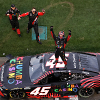Tyler Reddick stands on the roof of his race car with both fists raised in triumph as his crew members cheer on the ground below.