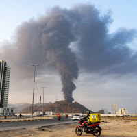 Delivery-persons ride motorcycles along a road as behind a tall smoke plume billows in the distance near a building