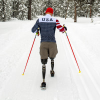A man with two prosthetic legs  walks in a snowy landscape using two red hiking poles.