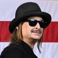 Kid Rock walks across a stage, military in the audience stand to clap, an American flag backdrop is behind them all