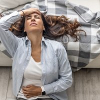 A woman sits on the floor in the living room, leaning her back against a white sofa, and holds her hands on her lower abdomen, suggesting that she is suffering from abdominal pain from menstruation or other health problems.