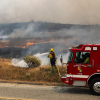 A firefighter standing in front of a fire truck aims a running hose at a fire overtaking a field by a road