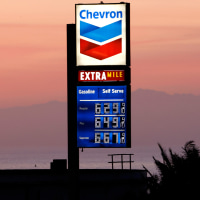 A Chevron gas station sign against a sunset sky with cars driving past.