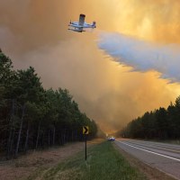 A plane drops a plume of white firefighting detergent over a tree line as the skies glow an orange color.