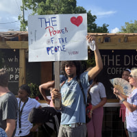 A young man holding a sign reading THE POWER OF THE PEOPLE at a small protest. 