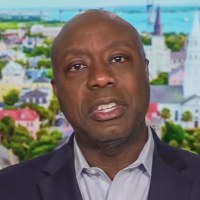 Tim Scott speaks while seated on set, a background of an aerial view of houses is behind him