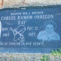 Carlos Ramon Obregon's headstone, flat on the ground, showing an engraving of Mother Mary on the left, a praying angel in the center, and Carlos on the right. The full headstone reads "Beloved Son & Brother, Carlos Ramon Obregon, "Ray", Nov. 13, 1963 - Dec. 12, 1977, You're forever in our hearts"