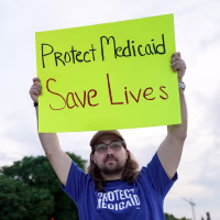 Two people hold signs outside, one the left it says "Medicaid funds critical disability services", and on the right it says "Protect Medicaid, Save lives"