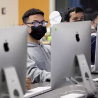 Students wearing facemasks sit behind a row of Apple desktop computers.