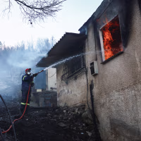 A firefighter sprays a hose into the window of a house on fire, in a charred forest environment