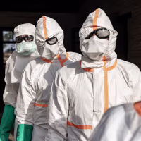 Several medical workers in white hazmat coveralls with masks and goggles standing in a line outdoors.