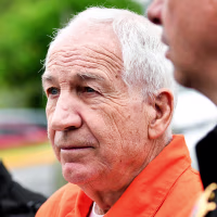 Jerry Sandusky holds paperwork. while walking in between two police officers outside, he wears an orange jumpsuit