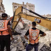 Two men wearing orange vests stand in front of two diggers in a field of debris.
