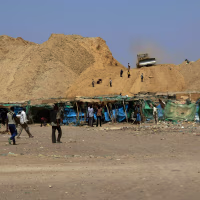Makeshift shelters lined up against a dry, dusty hillside with a front loader atop it.
