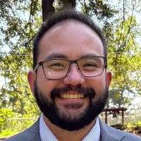Image: Jonathan McKinsey wearing a suit, stood in front of trees draped with string lights. 