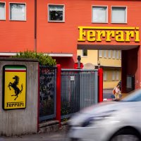 A detail view shows Ferrari factory signage, a yellow shield bearing the black prancing horse and Italian tricolor stripes, mounted on a concrete wall.