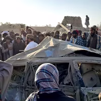 A crowd of men surround a charred and smashed vehicle.