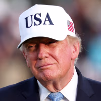 Donald Trump stands outside while wearing a white baseball cap that reads "USA" on the front