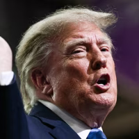 Donald Trump speaks at a podium, lifts one hand up in the air