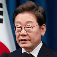 Lee Jae Myung speaks into a microphone, Korean flag can be seen behind him against a blue curtain