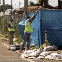 Workers in yellow vests removing blue tarp from a chainlink fence.