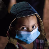 A child looks up at the camera wearing a blue facemask while standing outside