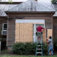 Two men place plywood over the windows of a home.