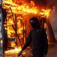 Image: A masked protester walks past a burning news-stand.