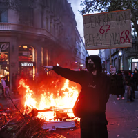 Image: A masked protestor holds a cardboard sign reading "Generations 6-7 redoes May 68" in front of a fire at the Boulevard des Capucines.