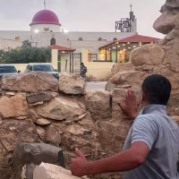 A man looks through a gap in a stone wall toward a Greek Orthodox church.
