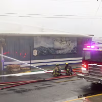 Two firefighters kneel in the street while spraying water on a burning building.
