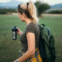 Woman is hiking in nature, with a backpack and holding a travel mug. The best travel mugs of 2021 include reusable cups, travel coffee mugs, ceramic travel mugs and stainless steel travel mugs from Ember, Contigo, YETI and more.